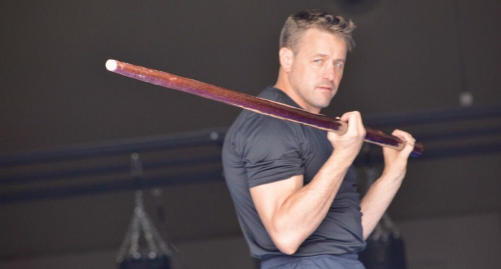 A man practicing with a Wing Chun dragon pole in a gym setting. He holds the long, tapered wooden staff in front of him with focus and attention, demonstrating a martial arts training exercise.