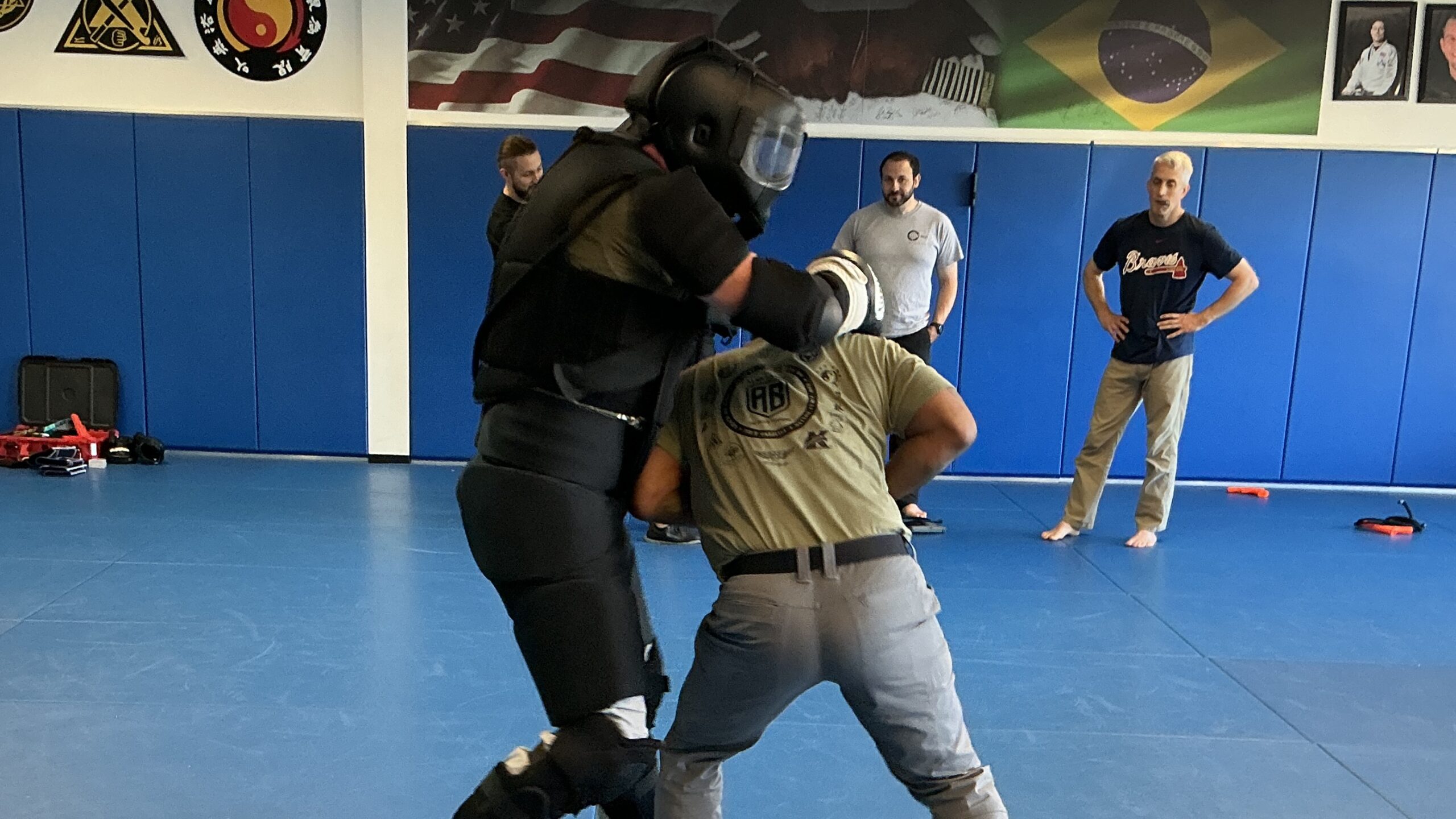 Two partners practice on blue mats, one wearing padded protective equipment, while other students observe.