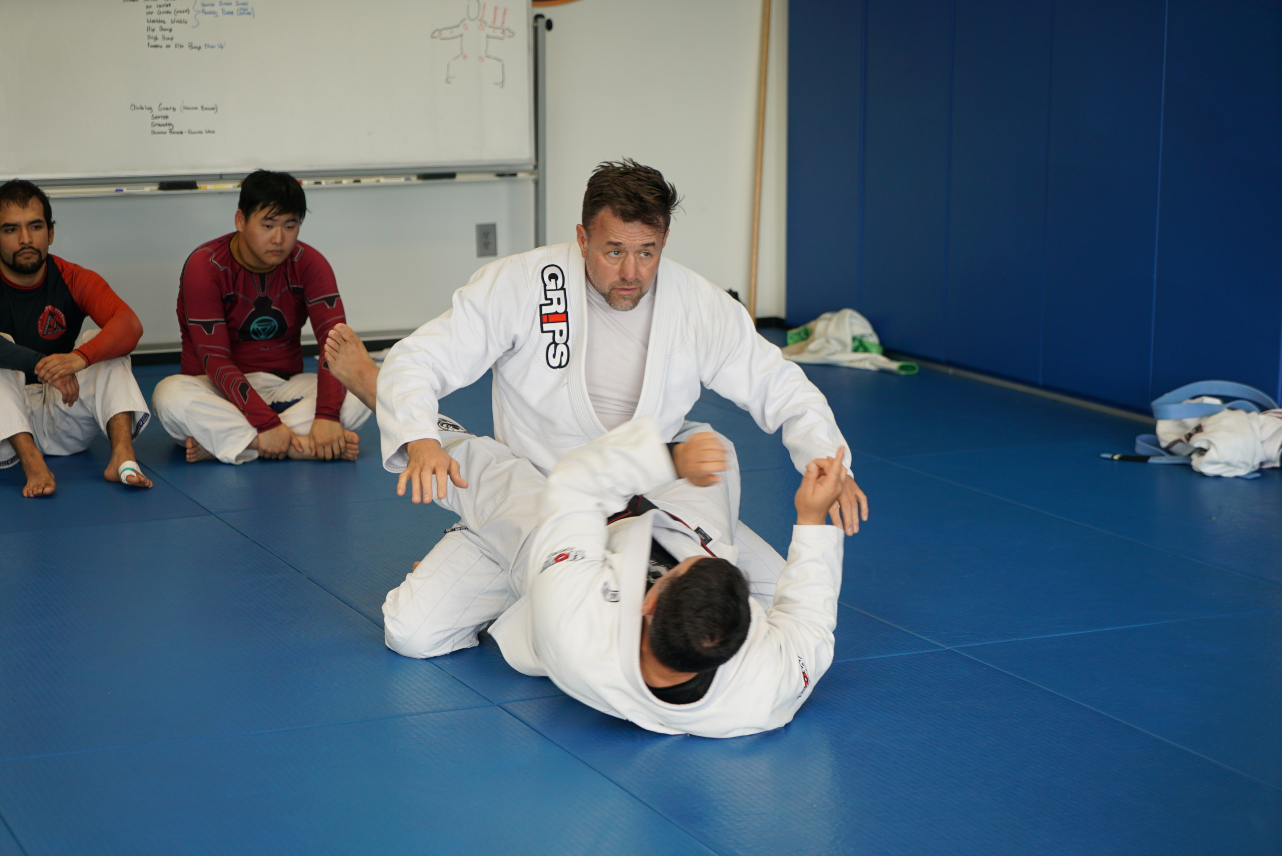 Alan Baker demonstrating Brazilian Jiu-Jitsu while students observe from the edge of the mat