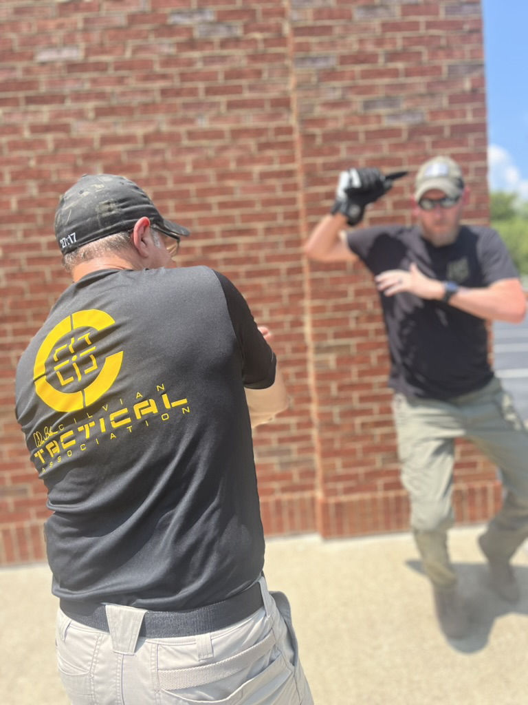 Two training partners facing one another beside a brick wall, with one holding a training implement overhead.